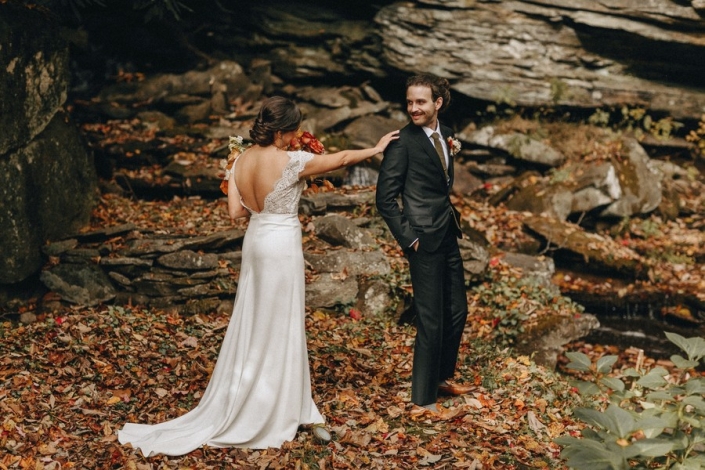 Bride taps groom on the shoulder during a first look on a rocky outcrop surrounded by autumn leaves.