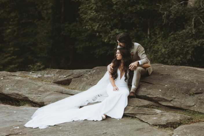 Newly married couple sit on a large rock formation together at a forest wedding venue.