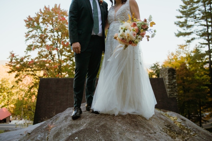 Bride and groom stand together on a large rock overlooking trees in fall color, the bride holding a colorful autumn bouquet.