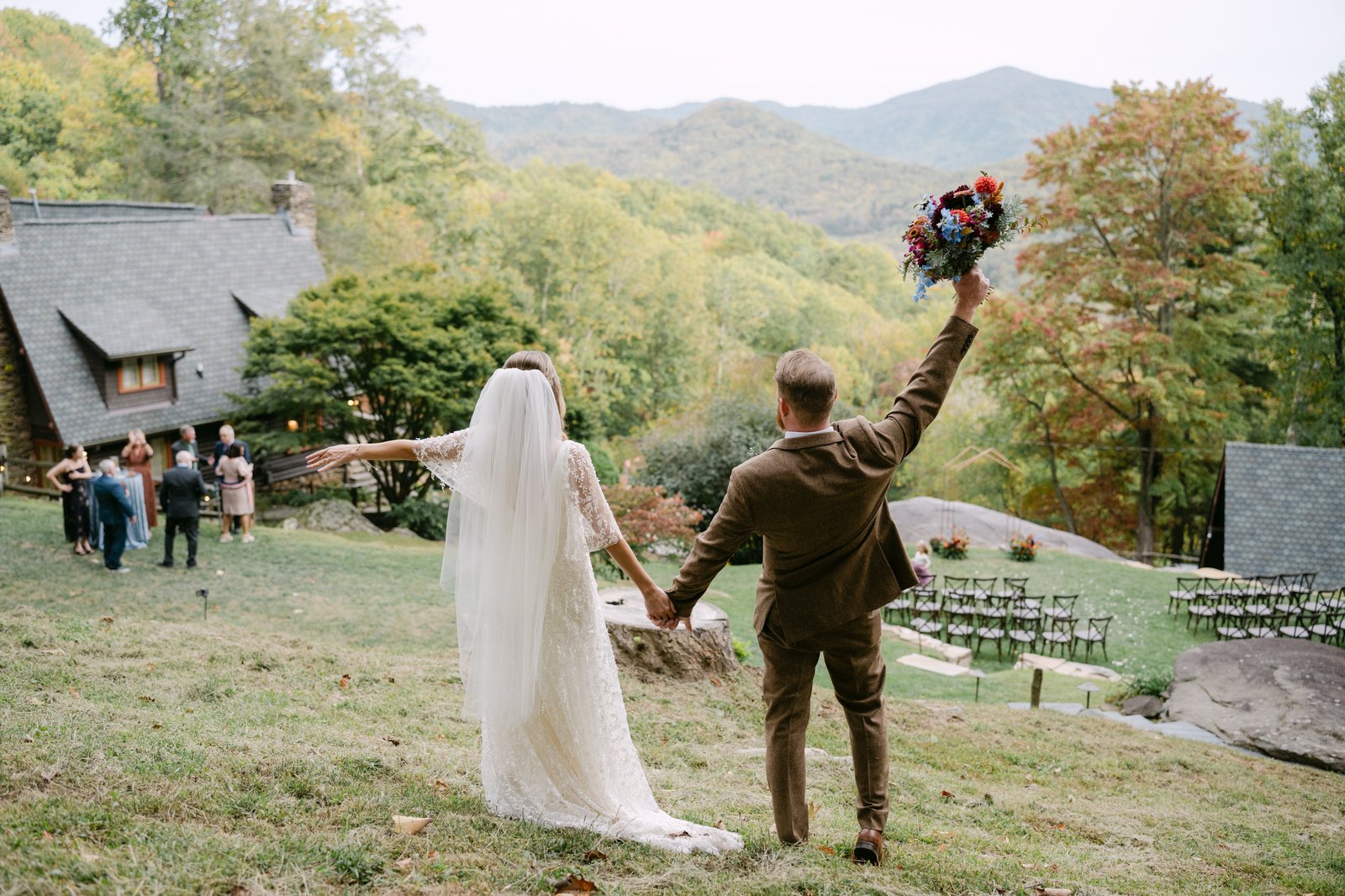 bride-and-groom-celebrate-overlooking-wedding-venue-laurel-falls-asheville-nc (1) Bride and groom celebrate with bouquet raised overlooking the Laurel Falls wedding venue, ceremony lawn, and Blue Ridge Mountains in fall