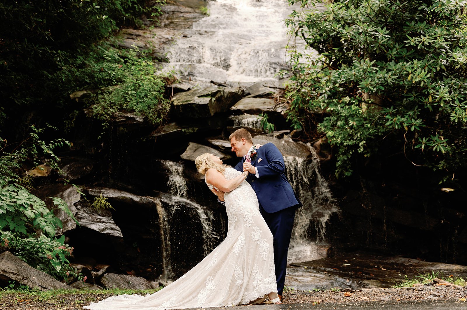 bride-and-groom-dip-kiss-waterfall-wedding-venue-laurel-falls-asheville-nc (1) Bride and groom embrace beside the on-site waterfall at Laurel Falls, a secluded mountain wedding venue near Asheville, NC