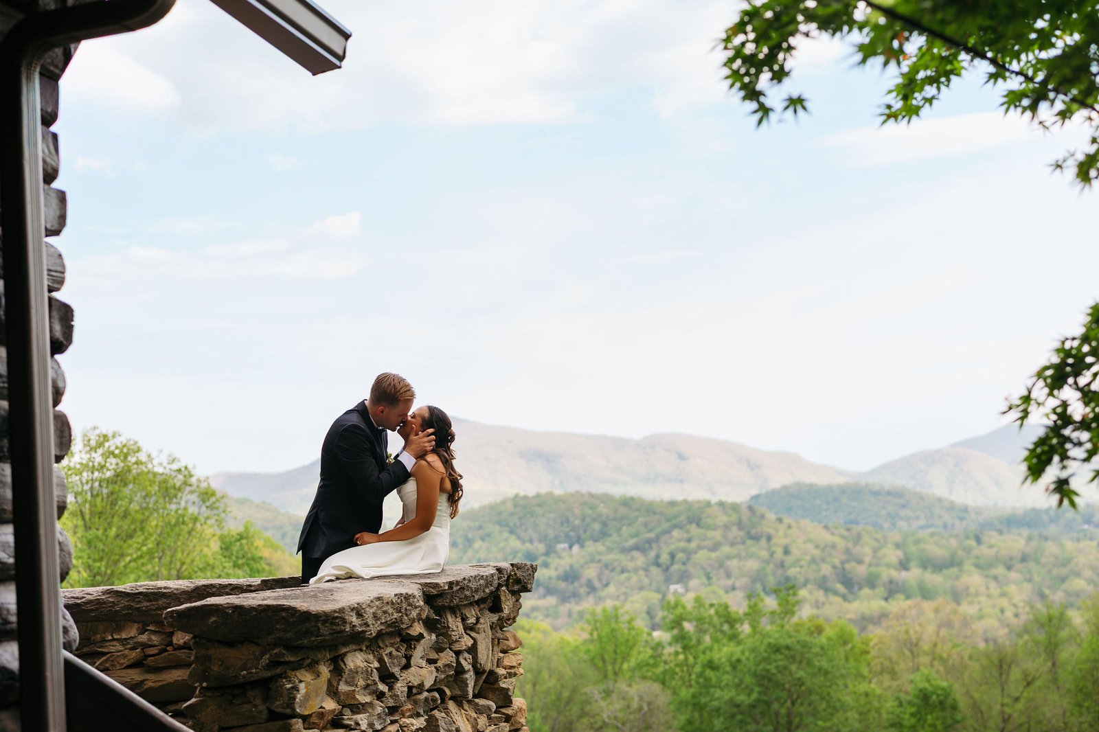 bride-and-groom-kiss-stone-wall-asheville-mountain-wedding-venue-laurel-falls (1) Bride and groom share a kiss on the stone wall overlook at Laurel Falls wedding venue with Blue Ridge Mountain views in the background
